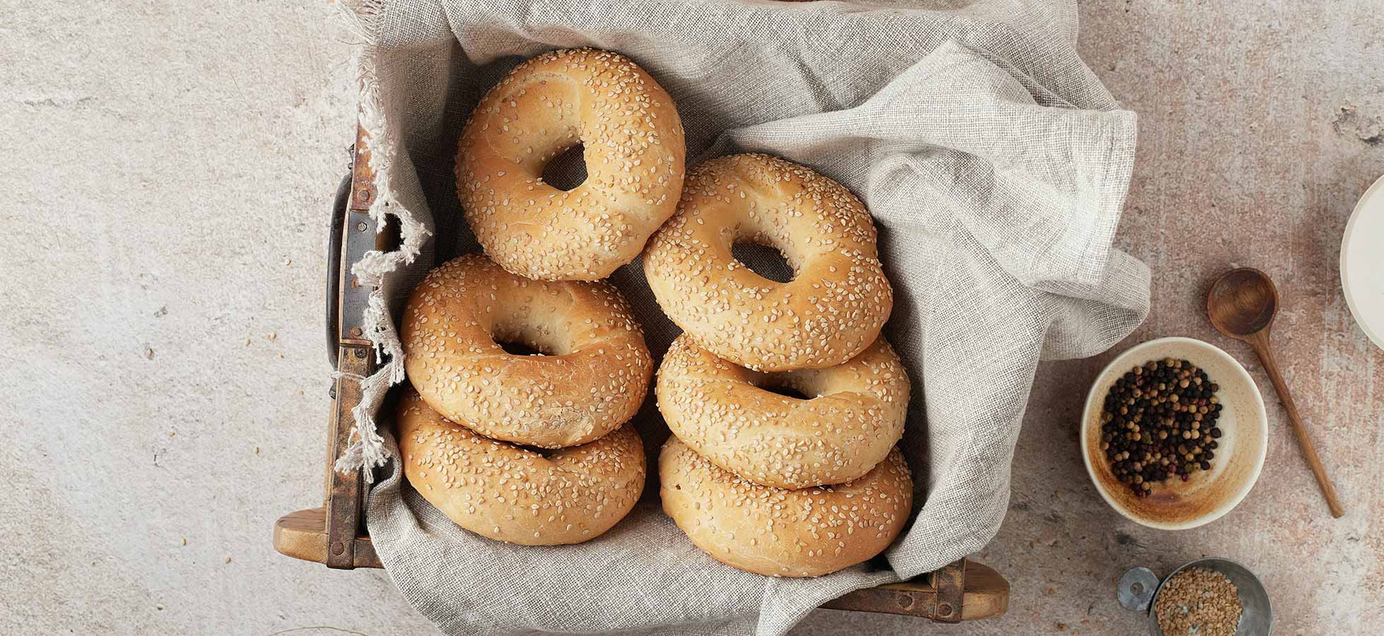 Ein Korb mit frisch gebackenen Sesam-Bagels, die ordentlich übereinander gestapelt sind. Im Hintergrund sind eine kleine Schüssel mit Gewürzen und ein Holzlöffel sichtbar. Die gesamte Anordnung vermittelt ein appetitliches und einladendes Gefühl.