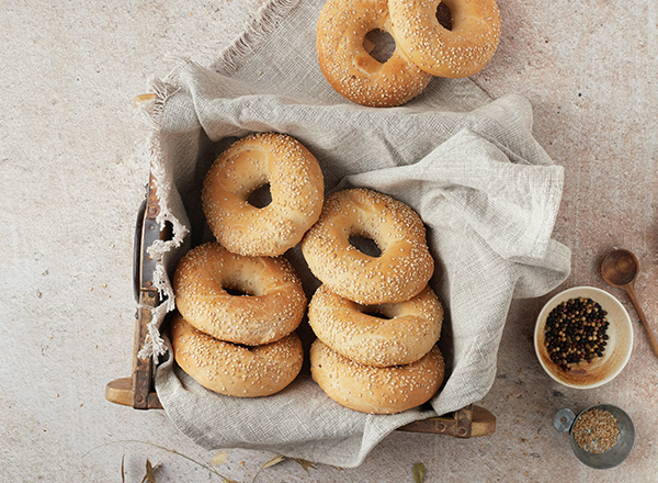 Ein Korb mit sieben frisch gebackenen Sesam-Bagels liegt auf einem Tisch, umgeben von einem grauen Stoff. Daneben befinden sich kleine Schalen mit verschiedenen Gewürzen. Die Bagels sind goldbraun und in unterschiedlichen Höhen angeordnet.