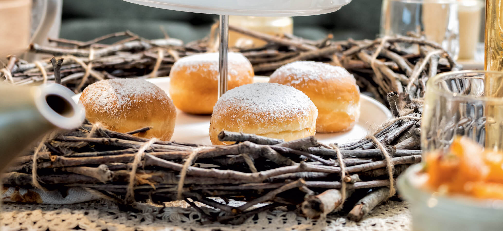 Auf einem dekorativen Teller liegen mehrere Berliner, die mit Puderzucker bestreut sind. Der Teller befindet sich in einem Kranz aus Zweigen, umgeben von weiteren Tafelelementen. Die Szenerie strahlt eine gemütliche Atmosphäre aus.