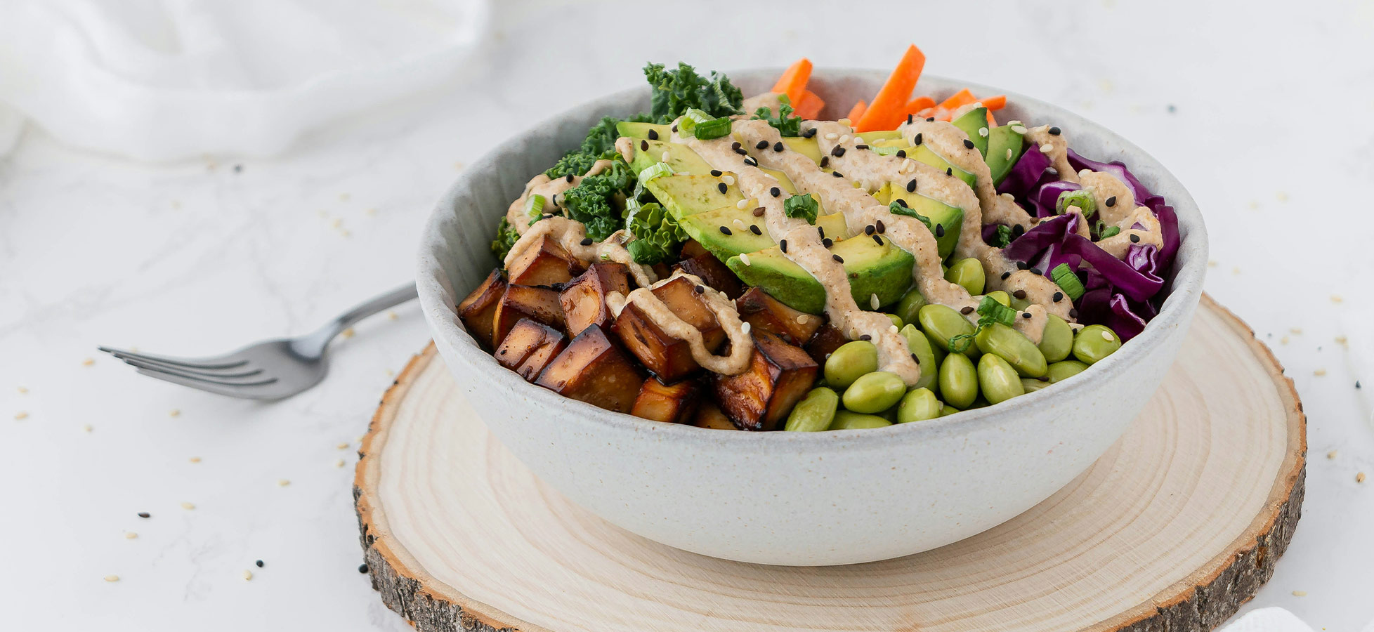 Eine Schüssel mit einer bunten Gemüseauswahl, darunter gebratener Tofu, grüne Edamame, lila Kohl, Avocado und Karottenstreifen, überzogen mit einer cremigen Sauce. Die Schüssel steht auf einem Holzbrett, umrahmt von einem hellen, dekorativen Hintergrund.