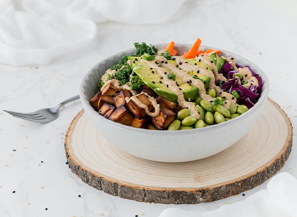Eine Schüssel mit einer bunten Gemüseauswahl, darunter gebratener Tofu, grüne Edamame, lila Kohl, Avocado und Karottenstreifen, überzogen mit einer cremigen Sauce. Die Schüssel steht auf einem Holzbrett, umrahmt von einem hellen, dekorativen Hintergrund.