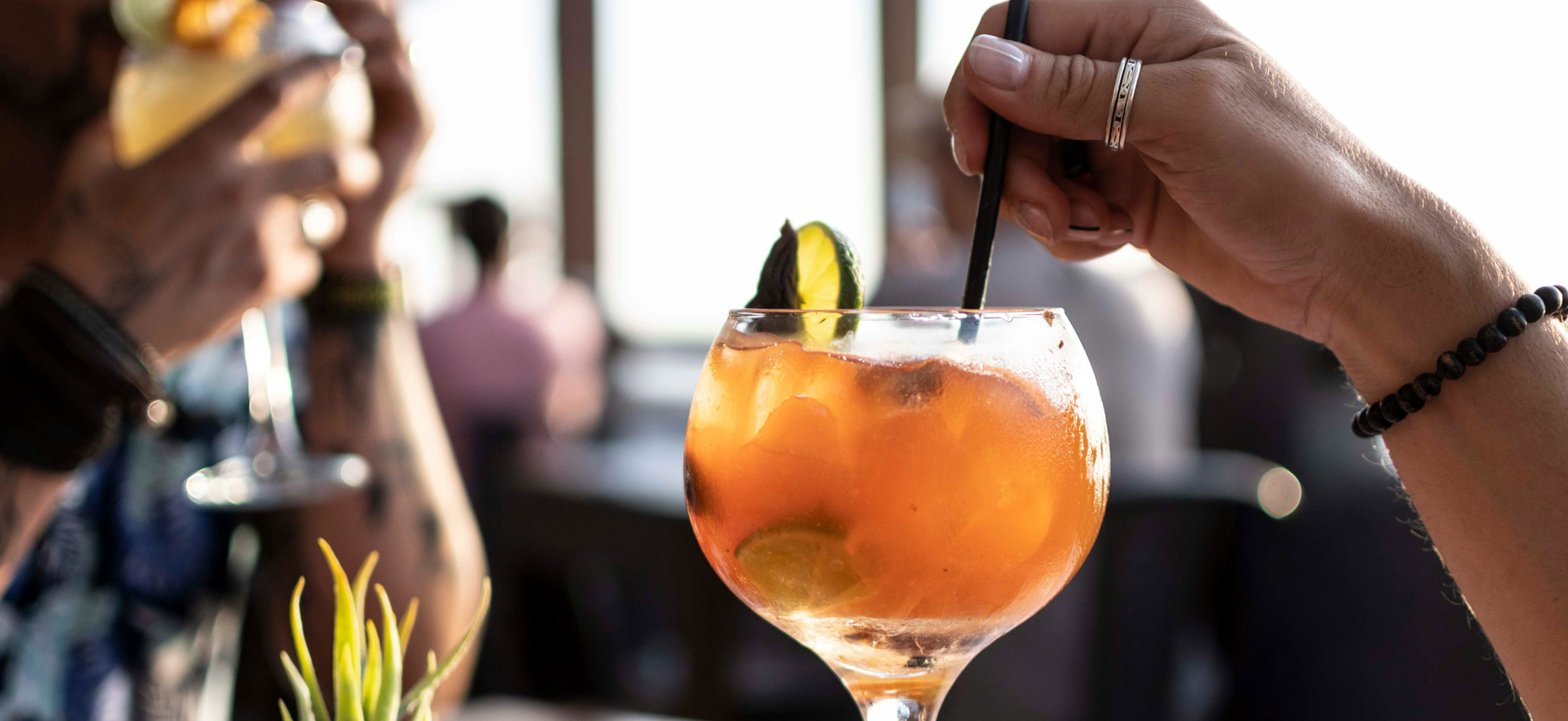 Ein fruchtiger Cocktail in einem großen Glas, gefüllt mit Eis und garniert mit einer Limettenscheibe und Minze. Eine Hand hält einen Strohhalm, während im Hintergrund eine unscharfe Silhouette einer weiteren Person zu sehen ist.