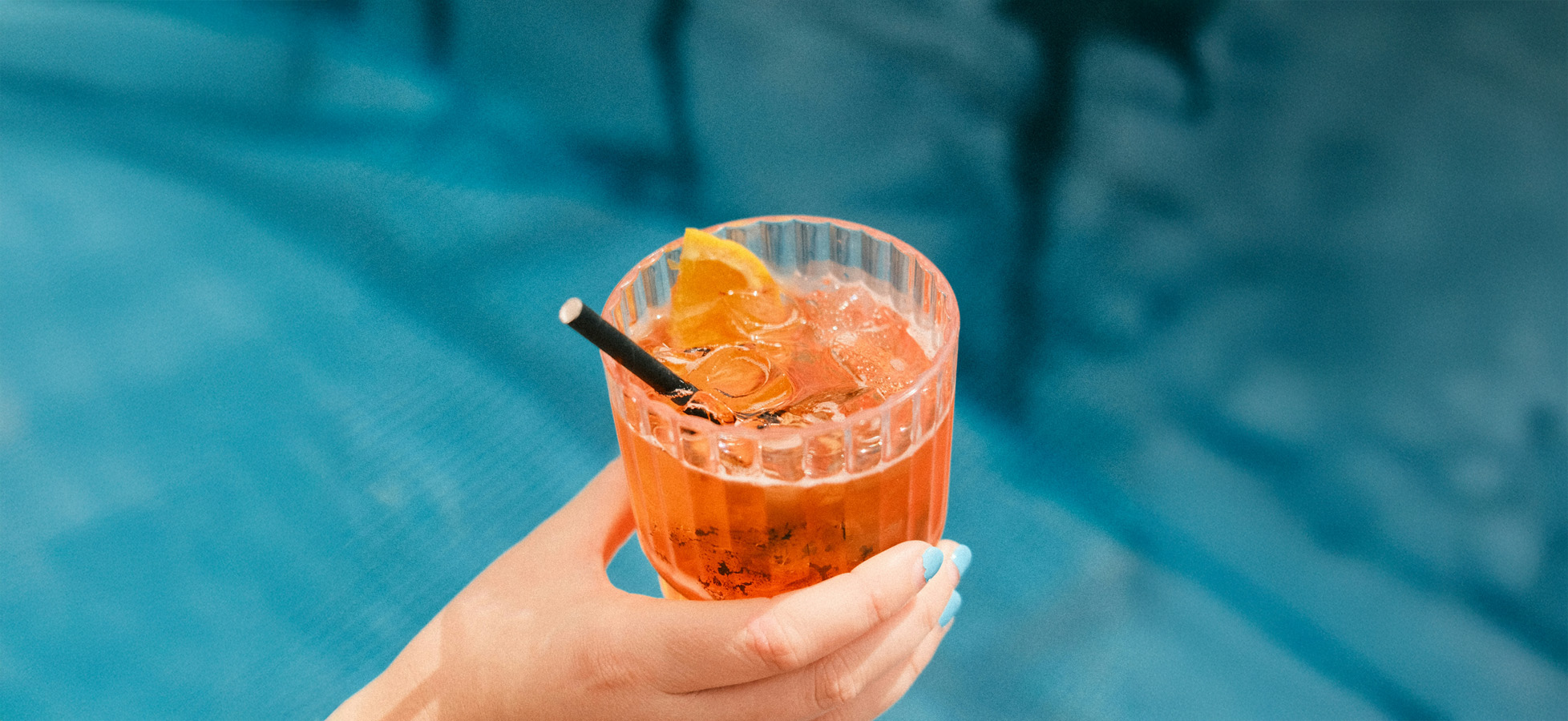 Ein handgehaltenes Glas mit einem bunten Getränk, das Eis und eine Scheibe Zitrone enthält. Im Hintergrund ist eine helle blaue Fläche zu sehen, die auf eine Poolumgebung hindeutet.