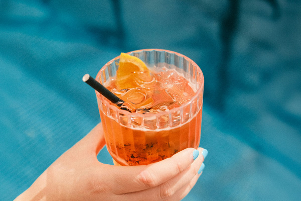 Ein Cocktailglas mit einem orangefarbenen Getränk, Eiswürfeln und einer Orangenscheibe dekoriert. Es wird von einer Hand gehalten, während der Hintergrund einen blauen Pool zeigt.