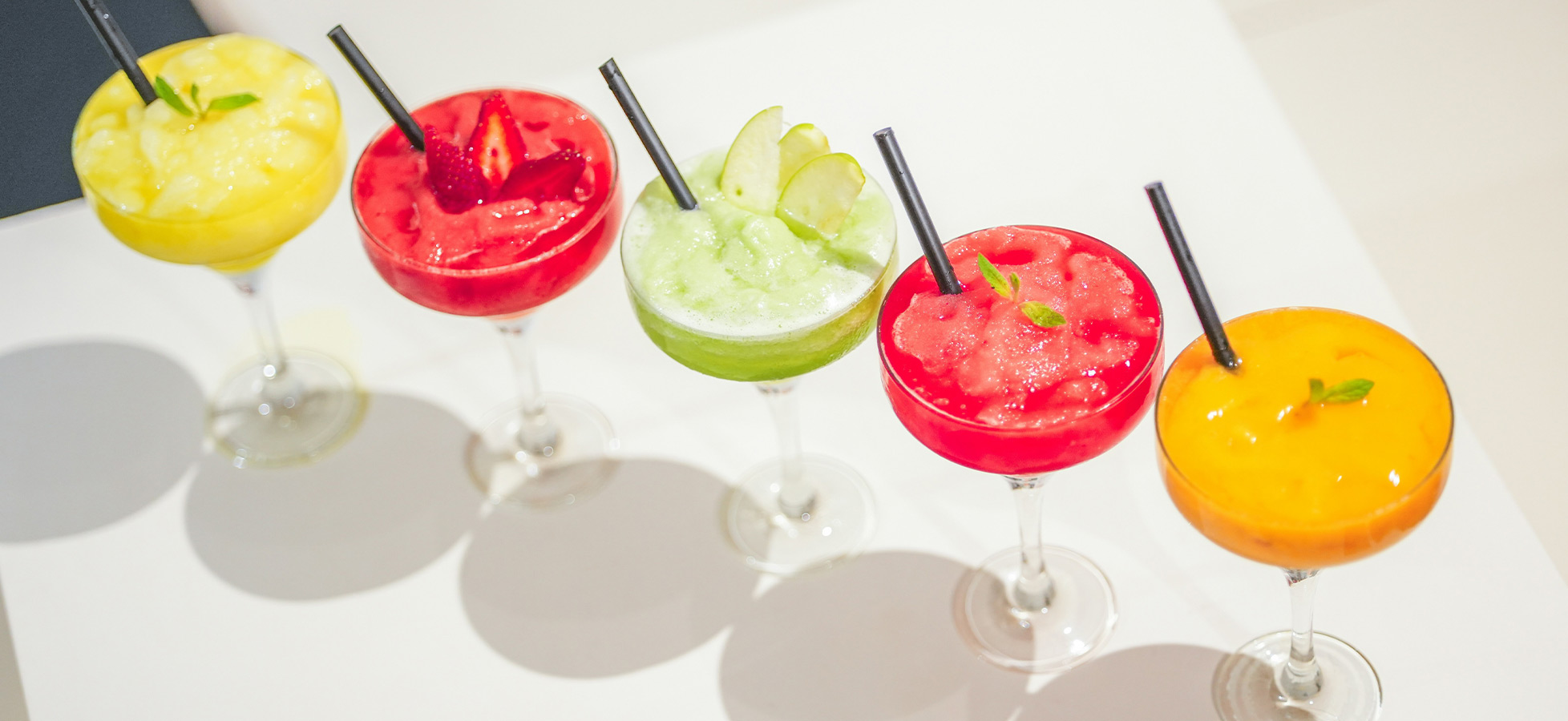 Fünf bunte Cocktails in eleganten Gläsern mit Schirmchen und Strohhalmen. Die Drinks haben verschiedene Farben: Gelb, Rot, Grün, Hellrot und Orange, und sind alle mit Eis versehen. Sie stehen nebeneinander auf einer weißen Unterlage.