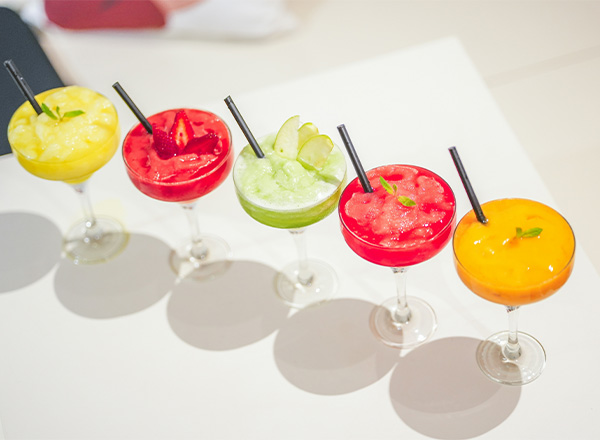 Fünf bunte Cocktails in eleganten Gläsern stehen nebeneinander. Die Getränke sind in den Farben Gelb, Rot, Grün und Orange gehalten und mit Strohhalmen und frischen Kräutern garniert. Sie vermitteln eine fröhliche und einladende Atmosphäre.