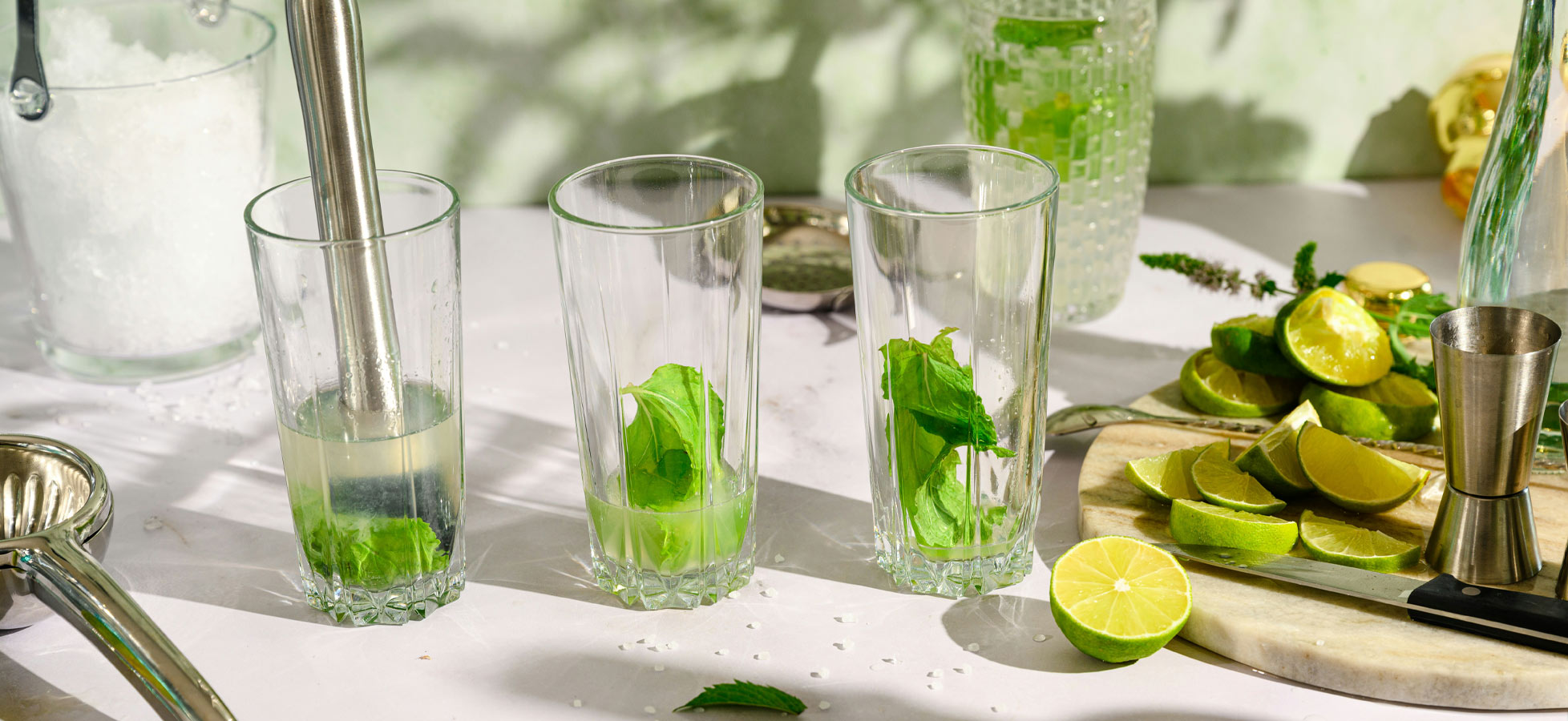 Drei Gläser mit frischen Minzblättern stehen auf einem Tisch, während Limettenscheiben daneben liegen. Im Hintergrund ist ein halb gefülltes Glas und weitere Zutaten sichtbar. Die Szene vermittelt eine erfrischende Getränkezubereitung, möglicherweise für einen Cocktaile.