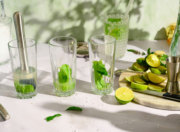 Drei Gläser mit frischen Minzblättern stehen auf einem Tisch, während Limettenscheiben daneben liegen. Im Hintergrund ist ein halb gefülltes Glas und weitere Zutaten sichtbar. Die Szene vermittelt eine erfrischende Getränkezubereitung, möglicherweise für einen Cocktaile.