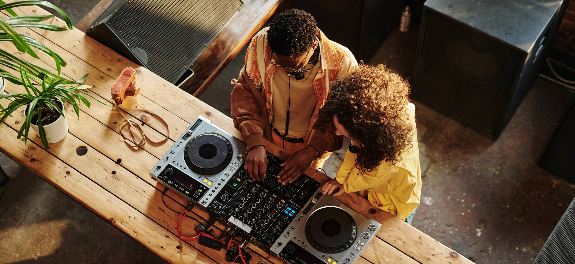 Zwei Personen stehen an einem Holztisch und bedienen DJ-Equipment. Eine Person mit lockigem Haar trägt ein gelbes Oberteil, während die andere in einem bunten Outfit gekleidet ist. Der Tisch ist mit Pflanzen und DJ-Geräten bedeckt, die in einem lebhaften Raum platziert sind.