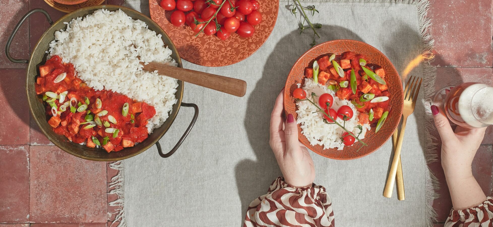 Eine Schüssel mit Reis und Gemüsegericht steht auf einem Tisch. Eine Person hält eine Tasse und hat eine weitere Schüssel mit Reis und Gemüse vor sich. Rote Kirschtomaten sind zusätzlich auf dem Tisch platziert. Der Tisch ist mit einem grauen Tuch bedeckt.