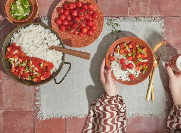 Eine vorbereitete Mahlzeit mit Reis und einer bunten Gemüse-Tomaten-Sauce ist auf einem Tisch angerichtet. Eine Hand hält eine Schüssel mit einer Portion des Gerichts, während eine andere Hand eine Gabel greift. Frische Tomaten und Frühlingszwiebeln sind ebenfalls sichtbar.