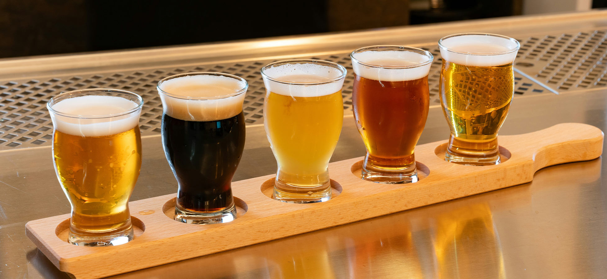 Das Bild zeigt eine Holzplatte mit fünf verschiedenen Biergläsern. Die Gläser enthalten Biere in unterschiedlichen Farben: von hellgelb bis dunkelbraun. Die Biere sind gleichmäßig angeordnet, was auf eine Verkostung hinweist. Der Hintergrund ist unscharf und lenkt nicht von den Gläsern ab.