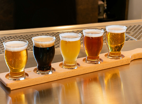 Fünf kleine Gläser mit verschiedenen Biersorten stehen auf einem Holzbrett. Die Gläser sind mit einer hellen und dunklen Flüssigkeit gefüllt, wobei jede Sorte eine unterschiedliche Farbe hat. Die Anordnung zeigt die Vielfalt der Biere.