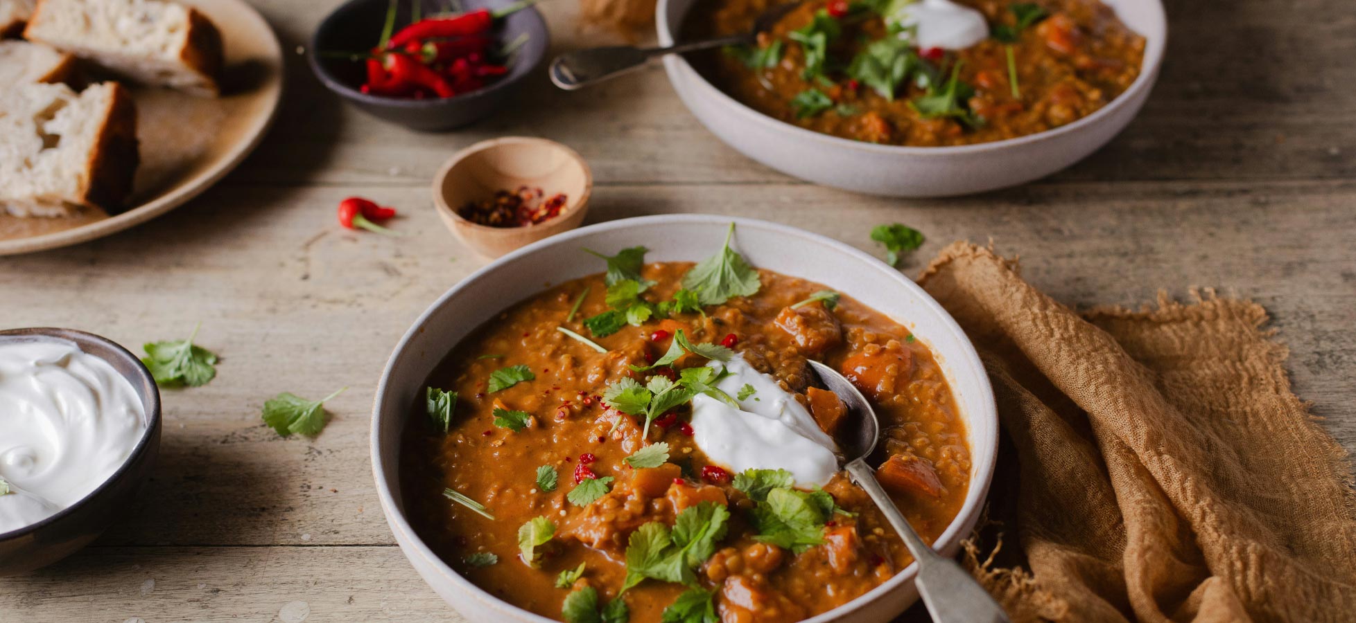 Eine Schüssel mit herzhaftem Eintopf, garniert mit frischem Koriander und einem Schuss Joghurt. Der Eintopf enthält verschiedene Gemüsestücke und Gewürze, während im Hintergrund eine weitere Schüssel sowie Brot und Chilis sichtbar sind.