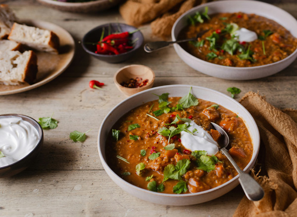 Zwei Schalen mit herzhafter Linsensuppe, garniert mit frischem Koriander und einem Klecks Joghurt. Dazu sind frisches Brot und rote Chilischoten auf einem Teller angerichtet. Der Hintergrund besteht aus einem rustikalen Holztisch.