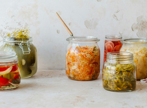 Mehrere Gläser mit fermentiertem Gemüse stehen auf einer hellen Oberfläche. Im Zentrum ein Glas mit orangefarbenem Sauerkraut, daneben Gläser mit unterschiedlichen eingelegten Gemüsearten. Einige Gläser sind transparent, sodass der Inhalt gut sichtbar ist.