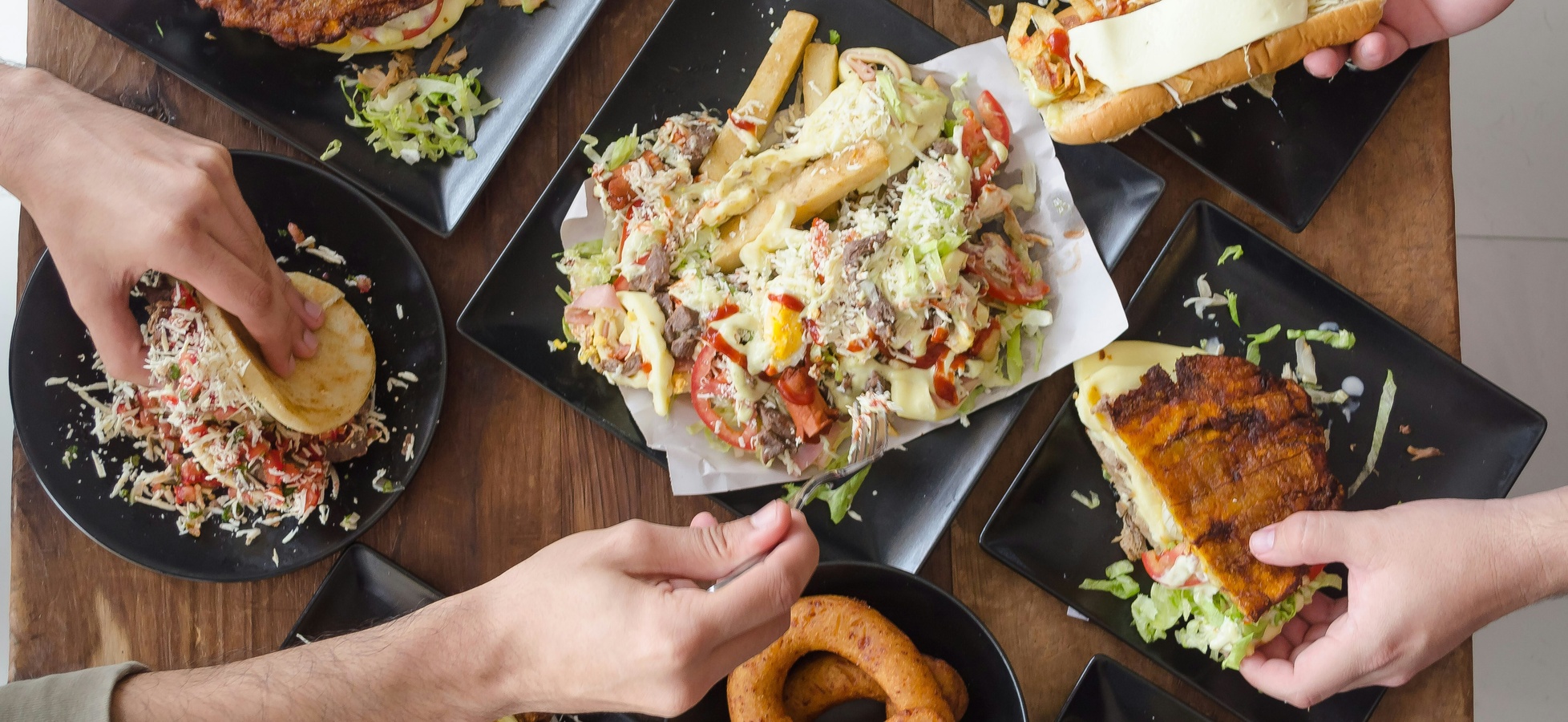 Mehrere Hände greifen nach unterschiedlichen Gerichten auf Tellern. In der Mitte befindet sich ein großer Teller mit Salat, Pommes und Fleisch, umgeben von weiteren Tellern mit Sandwiches und Snacks. Die Szene vermittelt eine gesellige Essensatmosphäre.