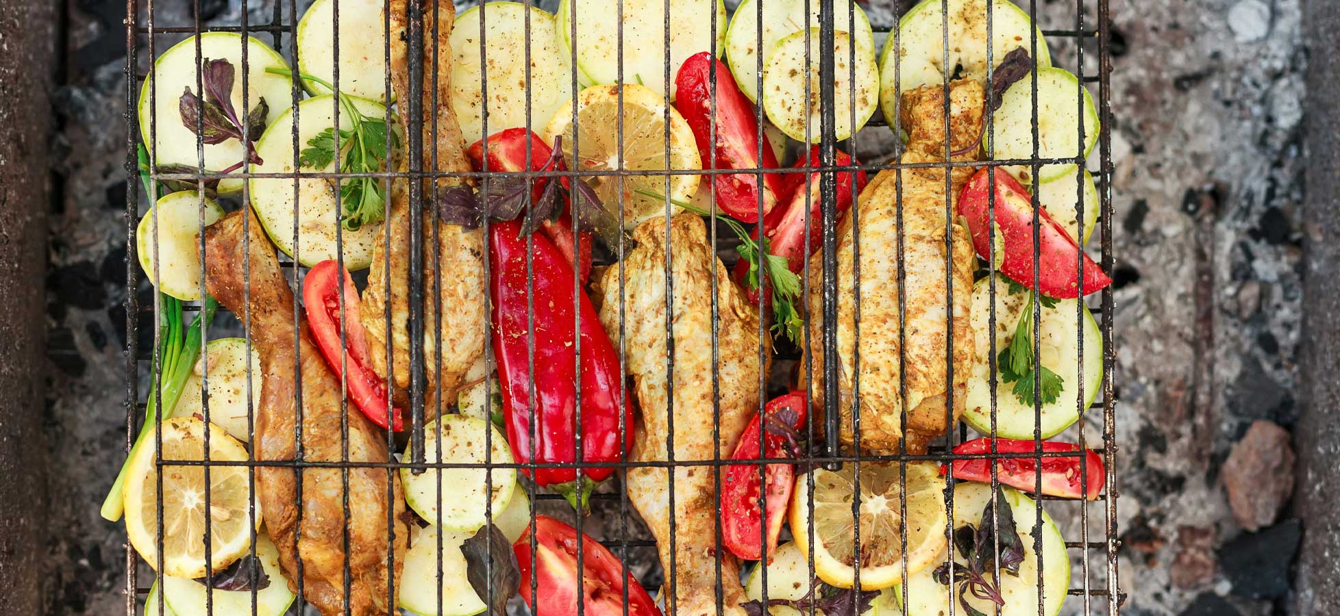 Ein Grill mit gegrilltem Hähnchen, umgeben von Zucchini, Paprika und Zitronenscheiben. Die Lebensmittel sind gleichmäßig auf einem Gitter angeordnet und zeigen eine appetitliche Mischung aus Gemüse und Fleisch. Der Grill ist über Kohlen angeordnet, was auf eine Outdoor-Kochsituation hindeutet.