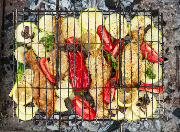 Ein Grillrost mit gegrillten Hühnchenschenkeln und verschiedenen Gemüsesorten, darunter Zucchini und Paprika. Alles ist gleichmäßig angeordnet und mit Kräutern garniert. Der Grillrost liegt auf einer Grillfläche mit glühenden Kohlen.