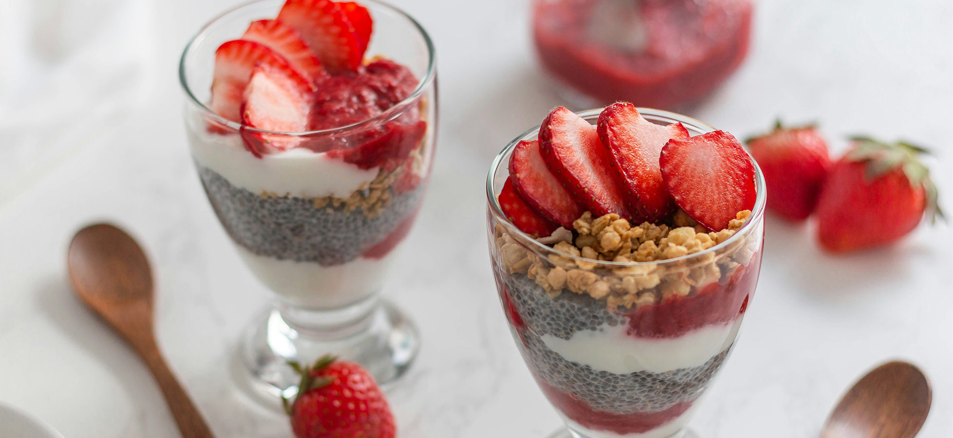 Zwei Gläser mit Schichtdessert sind auf einem Tisch präsentiert. Das Dessert besteht aus Joghurt, Chiasamen und Fruchtpüree, garniert mit frischen Erdbeeren und knusprigem Müsli. Rote Erdbeeren liegen ebenfalls auf dem Tisch. Ein Holzlöffel ergänzt das Arrangement.