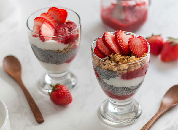 Zwei Gläser mit Schichtdessert sind auf einem Tisch präsentiert. Das Dessert besteht aus Joghurt, Chiasamen und Fruchtpüree, garniert mit frischen Erdbeeren und knusprigem Müsli. Rote Erdbeeren liegen ebenfalls auf dem Tisch. Ein Holzlöffel ergänzt das Arrangement.
