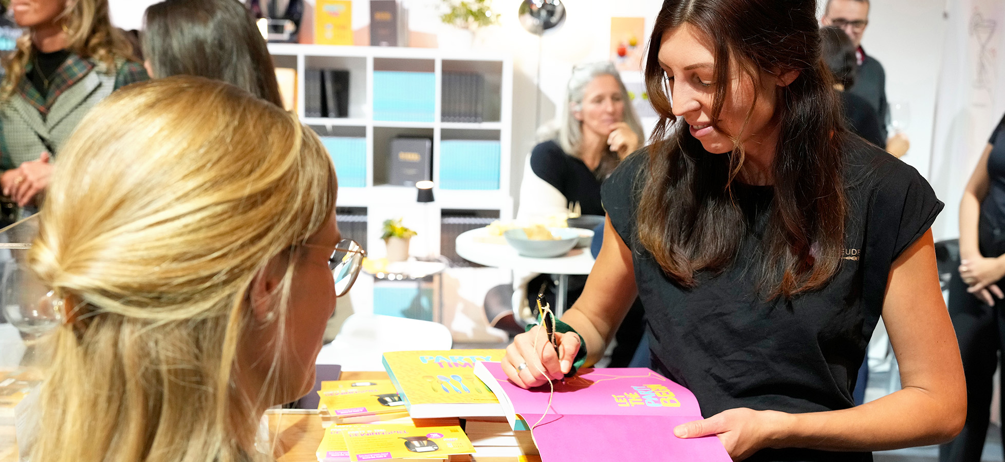 Eine Frau in schwarz signiert ein Buch für eine andere Person. Auf dem Tisch liegen weitere Bücher und Karten. Im Hintergrund sind weitere Personen zu sehen, die den Moment beobachten.