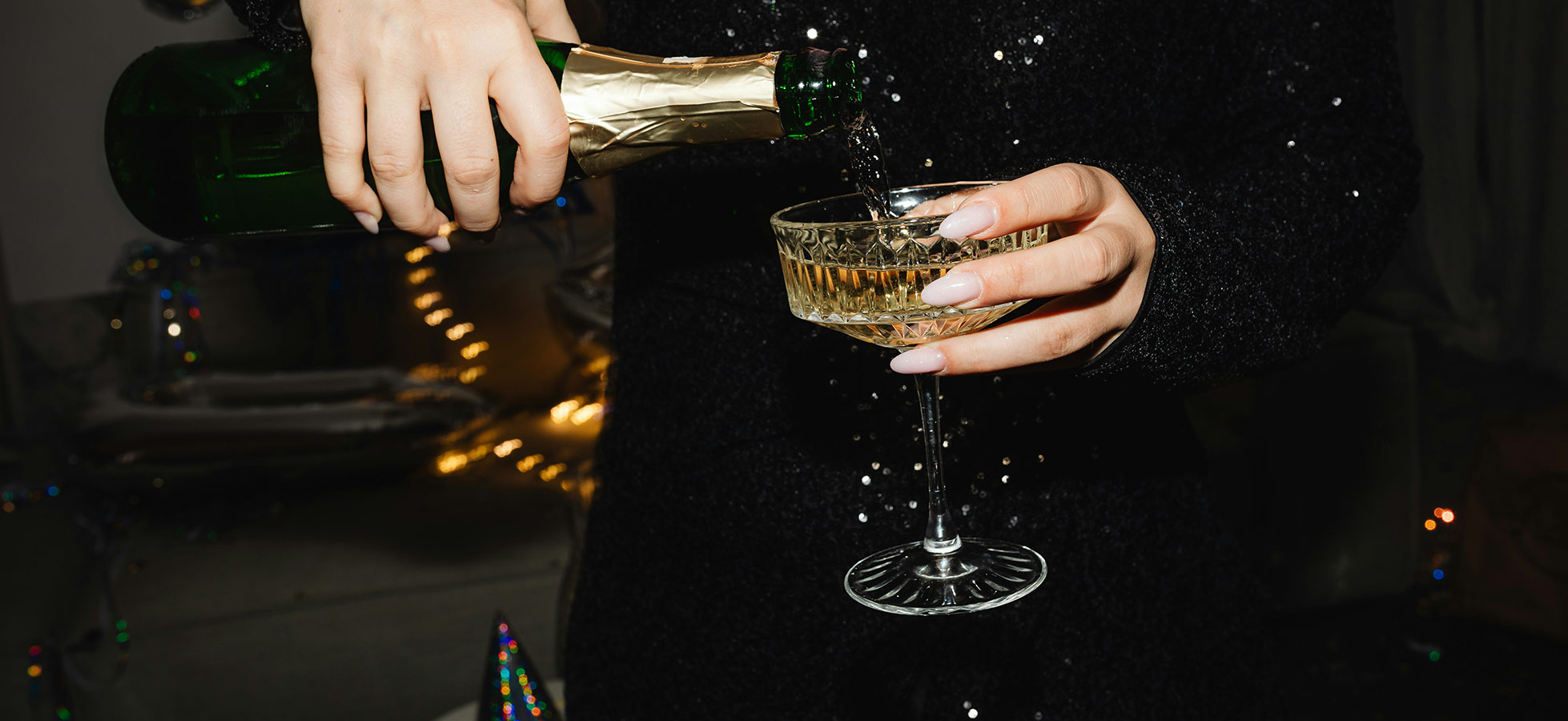 Eine Person in schwarzer Kleidung gießt Sekt aus einer grünen Flasche in ein elegantes Glas. Die Szene vermittelt eine festliche Atmosphäre, geeignet für Feierlichkeiten oder besondere Anlässe.