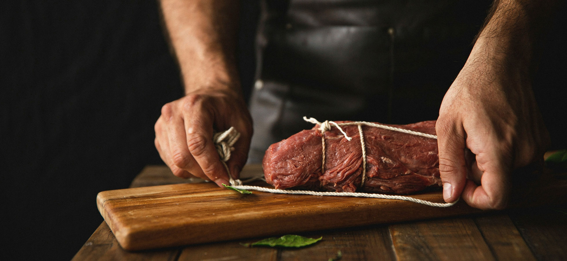 Ein Koch bindet ein Stück rohes Fleisch mit einer Schnur auf einem Holzbrett. Die Hände sind deutlich sichtbar und zeigen die Technik des Bindens. Im Hintergrund sind keine ablenkenden Elemente zu sehen.