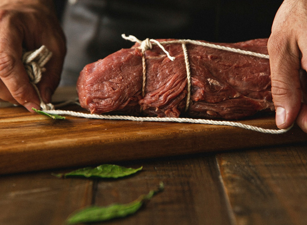 Ein rohes Stück Fleisch wird auf einem Holzbrett mit einer Schnur zusammengebunden. Dabei hält eine Hand das Fleisch, während die andere Hand die Schnur anlegt. Im Vordergrund sind einige grüne Blätter sichtbar.