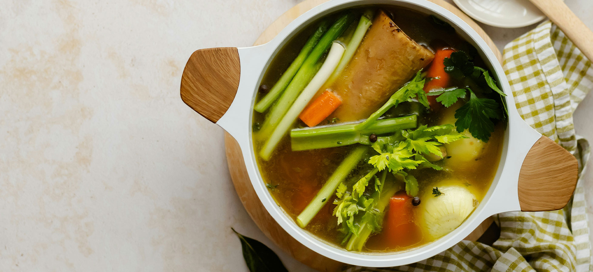 Eine weiße Schüssel mit leicht brühendem Gemüsebrühe ist im Bild zu sehen. Die Brühe enthält sellerie, Karotten, frische Kräuter und ein Stück Fleisch. Die Schüssel steht auf einem Holzbrett, umgeben von einem karierten Tuch.