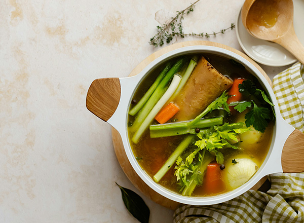 Ein weißer Suppentopf mit einer bunten Gemüsesuppe, die Sellerie, Karotten und Zwiebeln enthält. Der Topf steht auf einem Holzuntersetzer, umgeben von frischen Kräutern. Ein Holzlöffel und ein kleines, rundes Geschirr sind ebenfalls im Bild zu sehen.