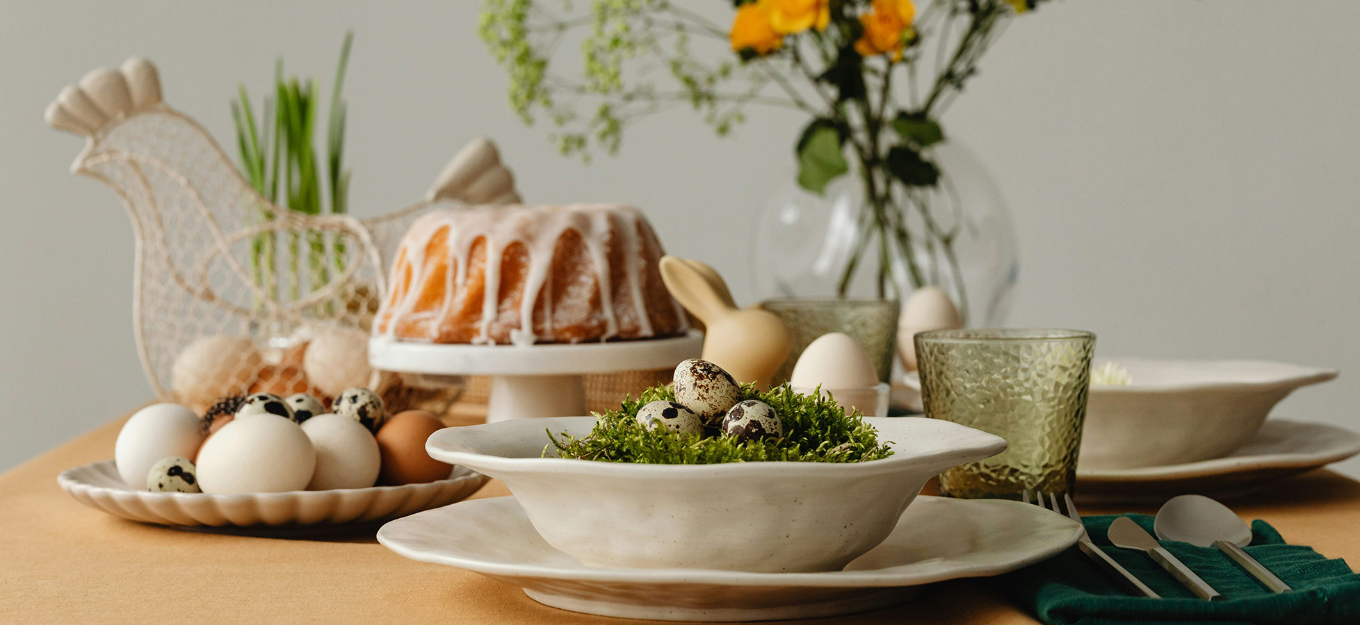 Ein gedeckter Tisch mit einer festlichen Anordnung. Im Mittelpunkt steht eine Schüssel mit bunten Eiern und frischem Grün. Daneben eine Torte auf einem Ständer, Gläser und Teller. Im Hintergrund ein Blumenstrauß und dekorative Elemente, die eine einladende Atmosphäre schaffen.