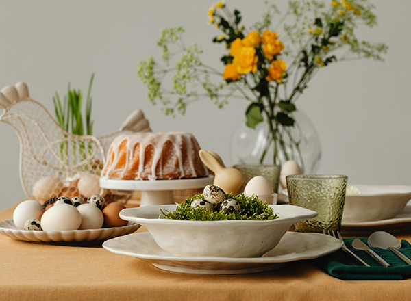 Ein festlich gedeckter Tisch mit verschiedenen Tellern und Schalen. Im Vordergrund liegen Eier auf einem Bett aus Grün, während im Hintergrund ein Gugelhupf und eine Blumenvase mit frischen Blumen sichtbar sind. Grüne Gläser und Besteck ergänzen die Dekoration.