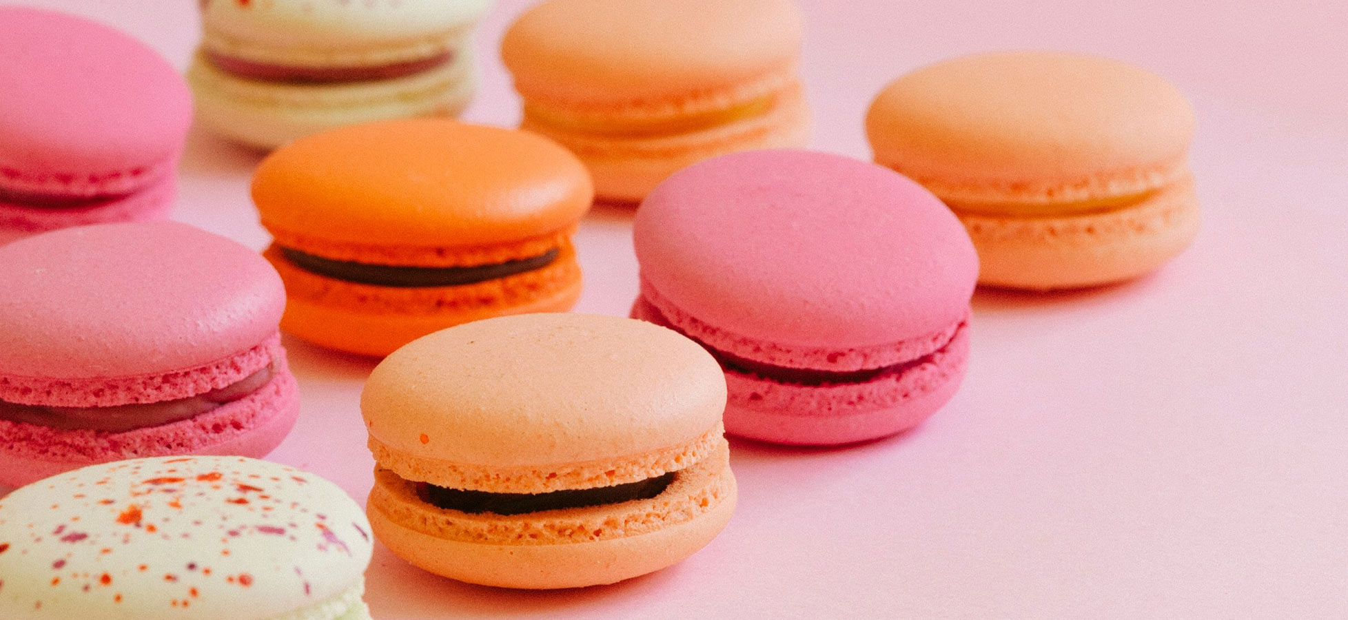 Eine bunte Auswahl an Macarons in verschiedenen Farben, darunter Pink, Orange und Gelb, steht auf einem rosa Hintergrund. Die Macarons sind rund und haben eine glatte Oberfläche. Sie befinden sich in unterschiedlichen Anordnungen in der Bildmitte.