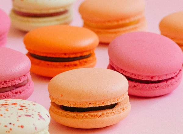 Eine Gruppe von bunten Makronen in verschiedenen Farben, darunter rosa, orange und grün, liegt auf einem rosa Hintergrund. Die Makronen sind rund, mit einer cremigen Füllung in der Mitte.