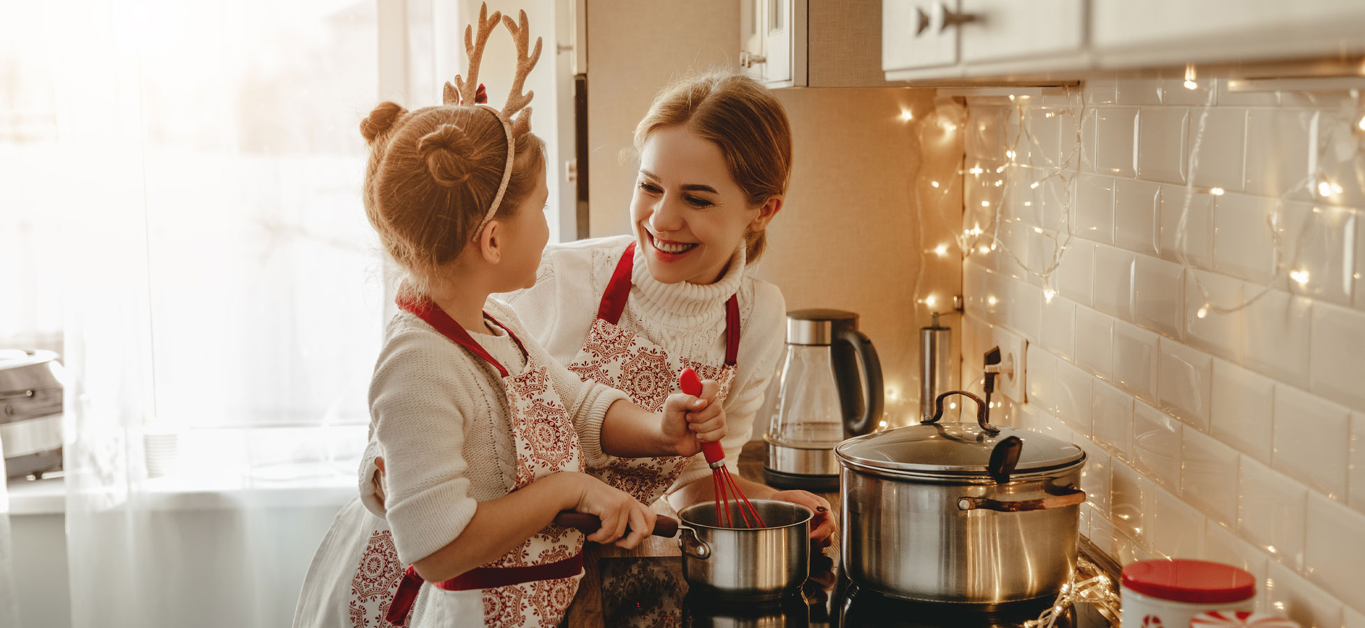 Eine Mutter und ihre Tochter stehen in einer hell erleuchteten Küche. Beide tragen Schürzen und lachen. Die Tochter hält einen Schneebesen, während die Mutter einen kleinen Kochtopf beobachtet. Die Atmosphäre ist freundlich und festlich, umrahmt von Lichtern im Hintergrund.