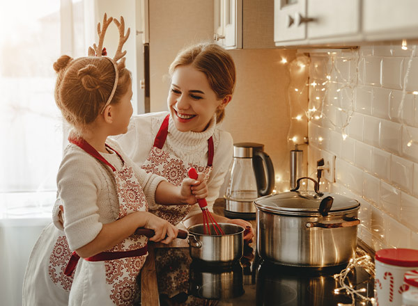 Eine Mutter und ihre Tochter stehen in einer hell erleuchteten Küche. Sie tragen festliche Schürzen und lächeln einander an, während sie zusammen in einem Topf rühren. Die Atmosphäre ist warm und einladend, mit sanften Lichtern im Hintergrund.