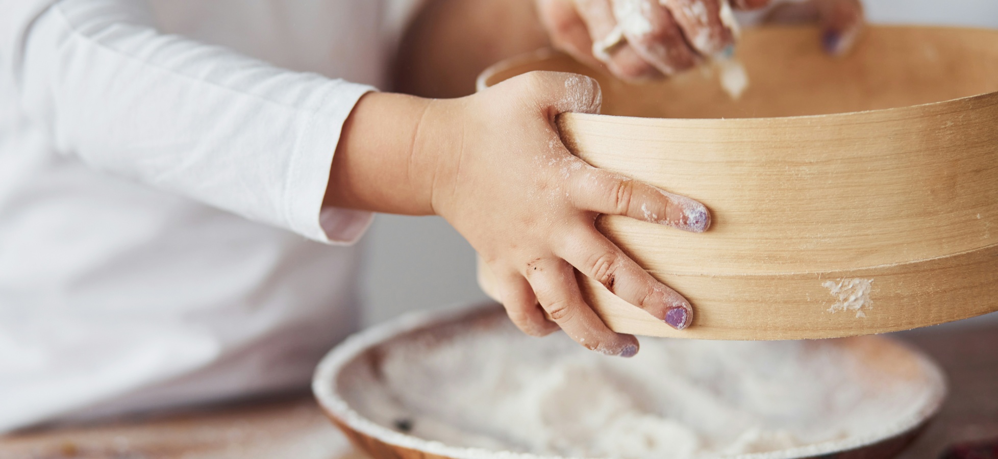 Ein Kind hält eine hölzerne Schüssel mit Teig oder Mehl in den Händen. Die Hände sind leicht mit Mehl bestäubt. Im Hintergrund steht eine andere Schüssel, die ebenfalls mit einer weißen Mischung gefüllt ist.