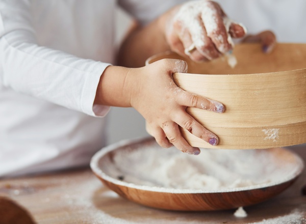 Ein Kind hält eine Holzschüssel mit Mehl, während eine andere Hand dabei hilft, die Zutaten zu mischen. Mehl ist auf der Arbeitsfläche verstreut, was auf das Backen hindeutet. Die Szene vermittelt eine gemütliche, familiäre Atmosphäre beim gemeinsamen Kochen.