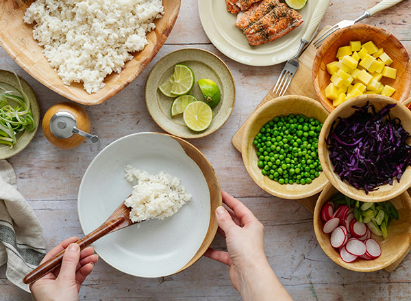 Eine Person ist dabei, gekochten Reis mit einem Löffel in eine weiße Schüssel zu schöpfen. Rund um die Person sind verschiedene Zutaten wie grüne Erbsen, rote Rettiche, Mango und Limetten in Schalen angeordnet. Im Hintergrund sind leicht gegrillter Lachs und frische Frühlingszwiebeln sichtbar.
