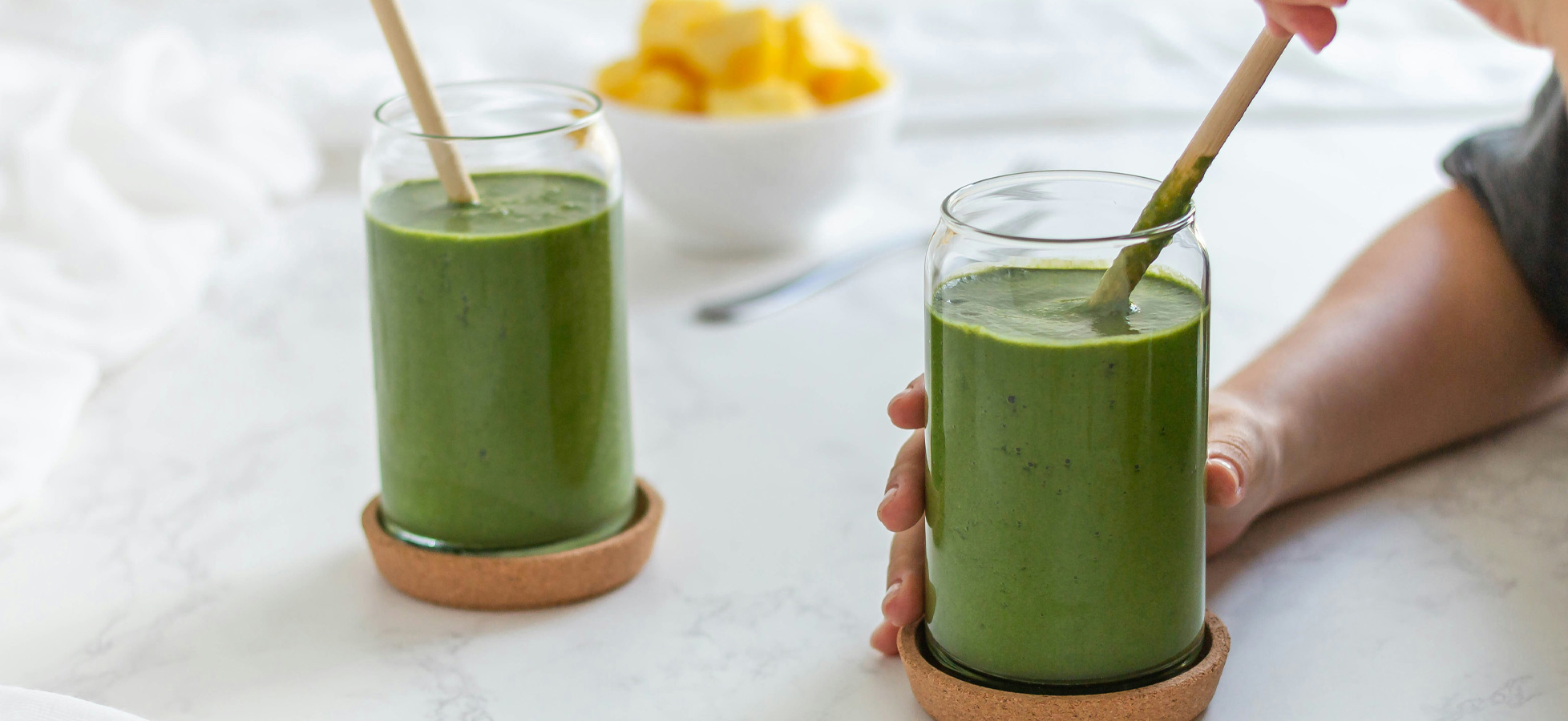 Zwei Gläser mit grünem Smoothie stehen auf einem Tisch. Eine Hand hält ein Glas mit einem Strohhalm, während ein weiteres Glas in der Nähe steht. Im Hintergrund befindet sich eine Schüssel mit gelben Früchten.