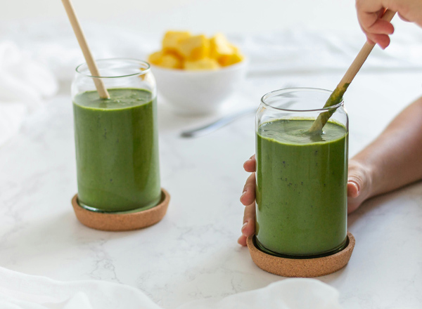 Zwei Gläser mit grünem Smoothie stehen auf einem Tisch. Eine Hand hält ein Glas mit einem Strohhalm, während ein weiteres Glas in der Nähe steht. Im Hintergrund befindet sich eine Schüssel mit gelben Früchten.