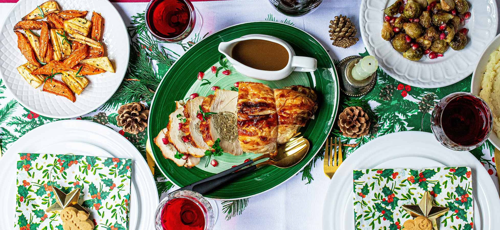 Ein festlich gedeckter Tisch mit einem Hauptgericht aus gebratenem Fleisch, umgeben von Beilagen wie geröstetem Gemüse und Kartoffelbrei. Rote Getränke sind in Gläsern angerichtet. Dekorative Servietten und Tannenzapfen verleihen der Szene eine weihnachtliche Atmosphäre.