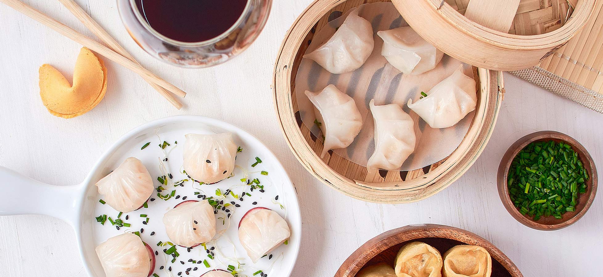 Ein Teller mit verschiedenen Teigtaschen, einschließlich gedämpfter Dumplings und frittierten Wontons, wird präsentiert. Dazu gibt es Soßen und Gewürze in kleinen Schalen. Im Hintergrund sind weitere Dumplings in einem Bambusdämpfer. Ein Glas Rotwein kompletziert das Bild.