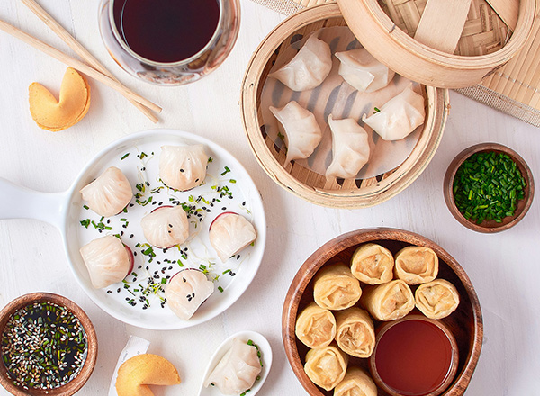 Ein Teller mit verschiedenen Teigtaschen, einschließlich gedämpfter Dumplings und frittierten Wontons, wird präsentiert. Dazu gibt es Soßen und Gewürze in kleinen Schalen. Im Hintergrund sind weitere Dumplings in einem Bambusdämpfer. Ein Glas Rotwein kompletziert das Bild.