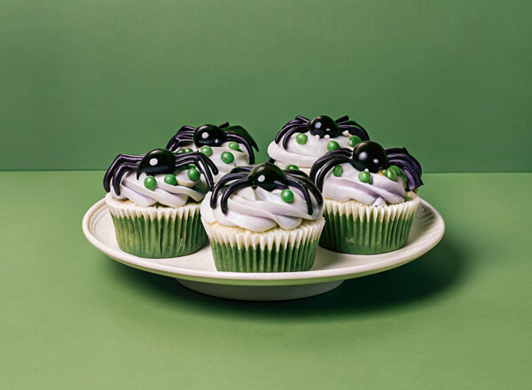 Sechs grüne Cupcakes auf einem weißen Teller mit lila Zuckerguss, verziert mit schwarzen Spinnen aus Süßigkeiten und grünen Streuseln. Der Hintergrund ist einfarbig grün.