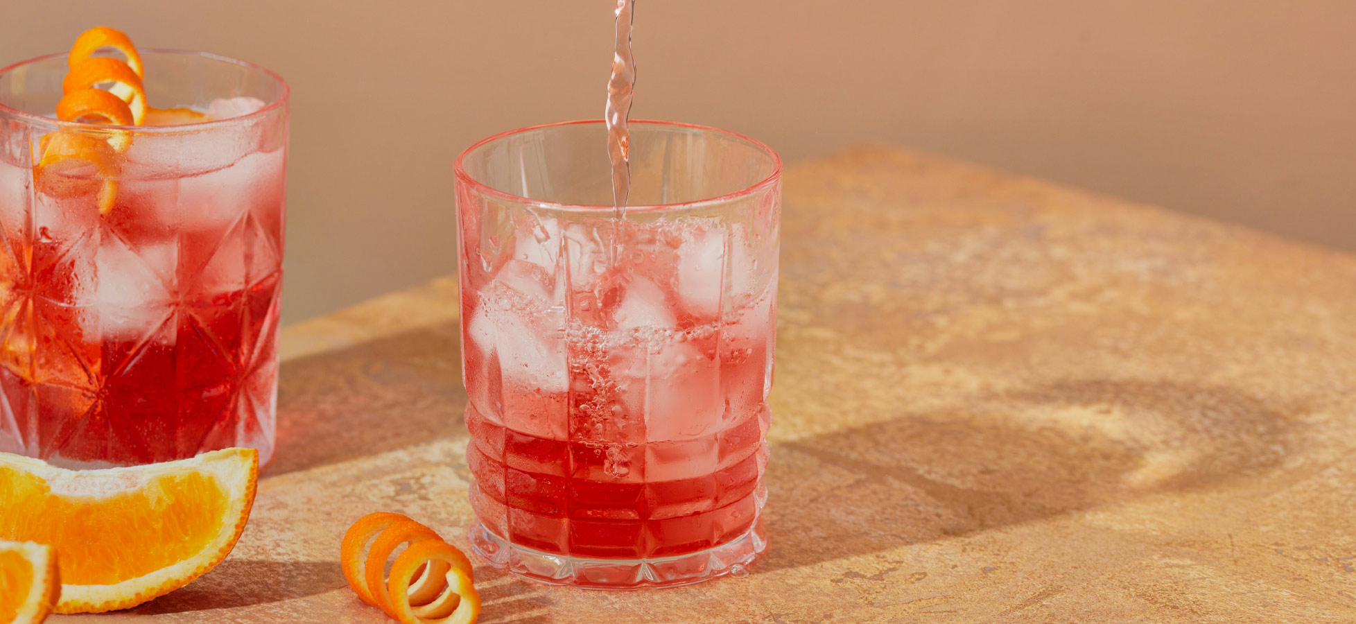 Zwei Gläser mit Eiswürfeln und roten Getränken stehen auf einem Tisch. Aus einem Kännchen wird frisches Wasser in das Glas auf der rechten Seite gegossen. Neben den Gläsern liegen Orangenscheiben und Zesten.