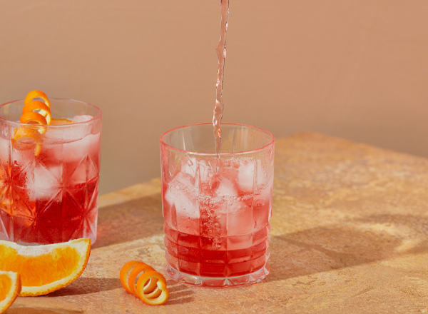 Zwei Gläser mit Eiswürfeln und roten Getränken stehen auf einem Tisch. Aus einem Kännchen wird frisches Wasser in das Glas auf der rechten Seite gegossen. Neben den Gläsern liegen Orangenscheiben und Zesten.