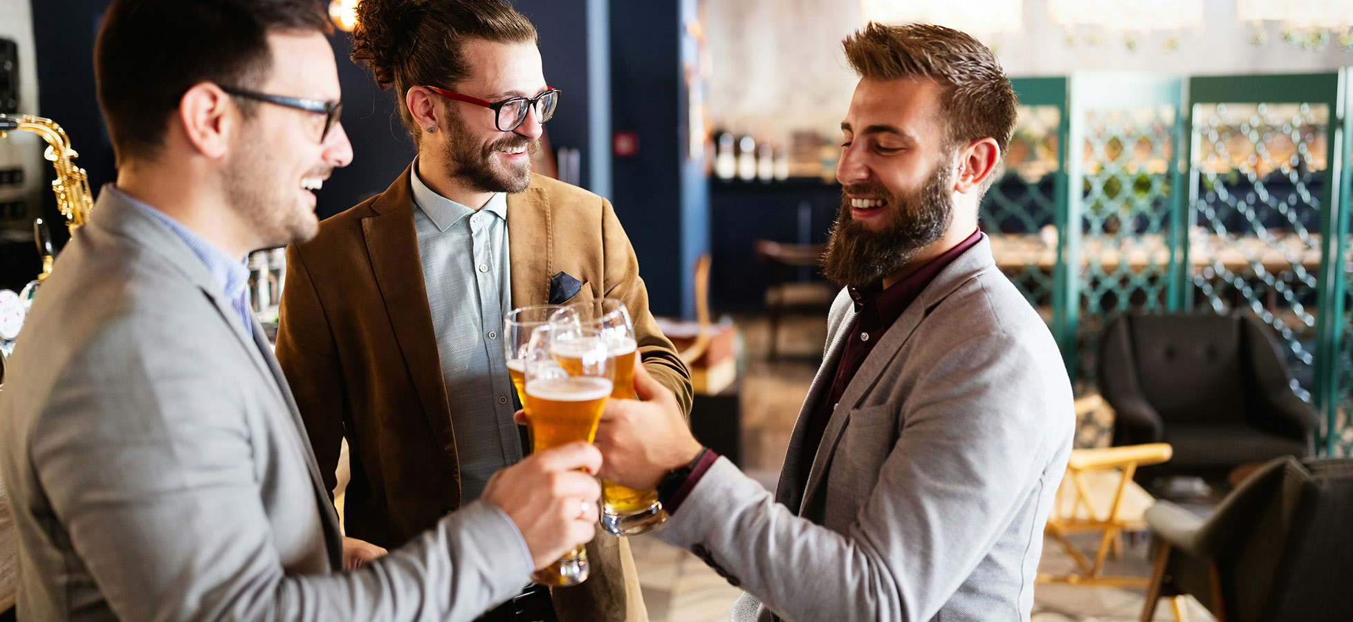 Drei Männer, gekleidet in Anzügen, stoßen mit Gläsern an, die Bier enthalten. Sie lächeln und scheinen eine fröhliche Zeit zu haben. Im Hintergrund ist eine moderne Bar zu sehen.