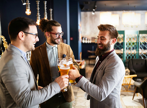 Drei Männer, gekleidet in Anzügen, stoßen mit Gläsern an, die Bier enthalten. Sie lächeln und scheinen eine fröhliche Zeit zu haben. Im Hintergrund ist eine moderne Bar zu sehen.